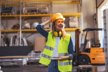 Görüş mesafesi yüksek giysiler içinde neşeli bir kadın işçi depoda bir pano tutuyor. Çalışmaya hazır, kendine güveni ve profesyonelliği gösteriyor, endüstri ve iş güvenliği duygusunu somutlaştırıyor..