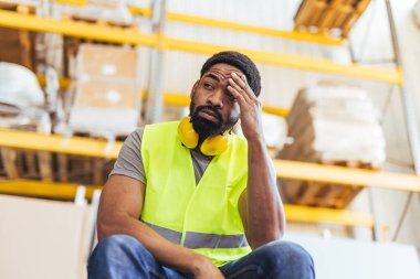 Koruyucu giysili bir depo çalışanı mola verirken yorulmuş görünüyor, yoğun iş gücü gerektiren işleri ve zorlukları yansıtıyor. Çalışma yorgunluğunu, işyeri koşullarını ve işçilerin refahının önemini vurgulamak.