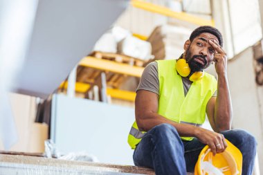 Yorgun bir inşaat işçisi otururken, yüksek görünürlüklü güvenlik donanımı giyerken ve elinde sıkı bir şapka tutarken, yorgunluk, yansıma ve endüstriyel işçiliğin talepkar doğasını yansıtır..
