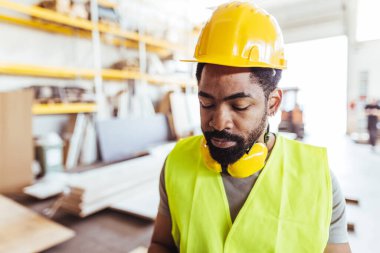 Depo ortamında koruyucu kask ve güvenlik yeleği giyen odaklanmış inşaat işçisi, bir çalışma sürecini veya durumunu dikkatle izliyor.