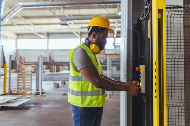 Sert şapka ve yansıtıcı yelek giyen bir sanayi işçisi, güvenlik ve verimliliği vurgulayarak fabrika ortamında makineler çalıştırıyor. Sahne profesyonellik ve endüstriyel otomasyon taşıyor.