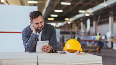 Profesyonel bir adam modern bir fabrikada tableti inceliyor. Yakınlarda, inşaat projeleri ve sanayi operasyonlarını vurgulayan sarı bir güvenlik kaskı bulunuyor. Resim, iş yeri planlamasını ve odaklanmasını gösterir.