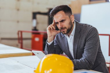 Resmi giysiler giyen bir mühendis, yakınlardaki bir çalışma alanında sabit bir şapkayla otururken, endüstriyel veya inşaat ortamında konsantrasyon ve sorun çözme işlemleri yaparken bir tablete odaklanır..
