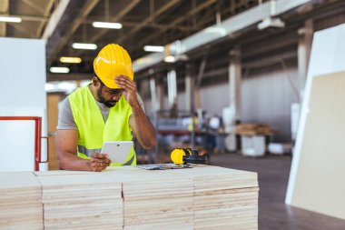 Güvenlik teçhizatı takan bir inşaat işçisi depoda dijital tablete danışıyor. Görüntü, teknoloji ve işyeri güvenlik tedbirlerine odaklı yoğun bir sanayi ortamını gösteriyor.