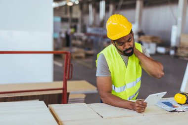 Elinde tablet tutan ve endişeli görünen bir inşaat işçisinin olduğu bir atölye sahnesi. Endüstriyel arka plan güvenlik ve teknolojik entegrasyon atmosferini artırıyor.