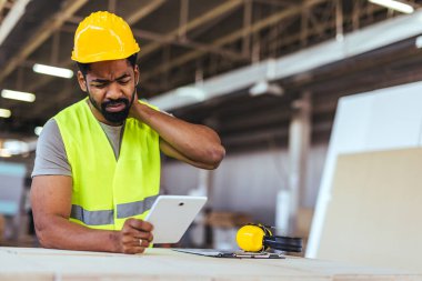 Başlık ve yelek giyen bir inşaat işçisi boyun rahatsızlığı hissederken tablet kullanır. Dekor ekipman ve doğal ışıklandırma ile dolu bir atölye..