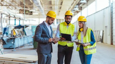 Çeşitli fabrika çalışanlarından oluşan bir ekip ve bir üretim tesisi içinde takım çalışması, liderlik ve endüstriyel güvenliği temsil eden, kask ve yüksek görünürlüklü yelek gibi koruyucu ekipmanlar giyen bir yönetici..