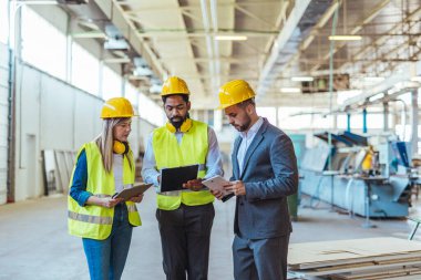 Çeşitli fabrika çalışanlarından oluşan bir ekip ve bir üretim tesisi içinde takım çalışması, liderlik ve endüstriyel güvenliği temsil eden, kask ve yüksek görünürlüklü yelek gibi koruyucu ekipmanlar giyen bir yönetici..