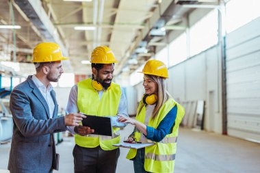 Çeşitli fabrika çalışanlarından oluşan bir ekip ve bir üretim tesisi içinde takım çalışması, liderlik ve endüstriyel güvenliği temsil eden, kask ve yüksek görünürlüklü yelek gibi koruyucu ekipmanlar giyen bir yönetici..