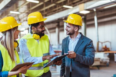 Çeşitli fabrika çalışanlarından oluşan bir ekip ve bir üretim tesisi içinde takım çalışması, liderlik ve endüstriyel güvenliği temsil eden, kask ve yüksek görünürlüklü yelek gibi koruyucu ekipmanlar giyen bir yönetici..