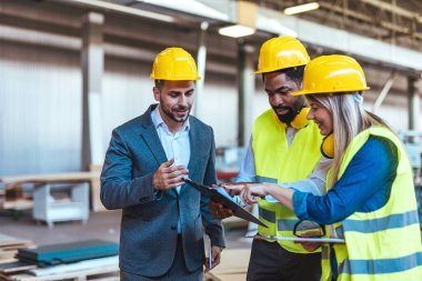 Çeşitli fabrika çalışanlarından oluşan bir ekip ve bir üretim tesisi içinde takım çalışması, liderlik ve endüstriyel güvenliği temsil eden, kask ve yüksek görünürlüklü yelek gibi koruyucu ekipmanlar giyen bir yönetici..