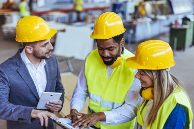 Çeşitli fabrika çalışanlarından oluşan bir ekip ve bir üretim tesisi içinde takım çalışması, liderlik ve endüstriyel güvenliği temsil eden, kask ve yüksek görünürlüklü yelek gibi koruyucu ekipmanlar giyen bir yönetici..