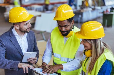 Çeşitli fabrika çalışanlarından oluşan bir ekip ve bir üretim tesisi içinde takım çalışması, liderlik ve endüstriyel güvenliği temsil eden, kask ve yüksek görünürlüklü yelek gibi koruyucu ekipmanlar giyen bir yönetici..