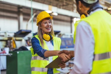 Fabrikada güvenlik donanımlı işçiler arasında dostça bir tokalaşma, endüstriyel çalışma ortamında takım çalışması, işbirliği ve profesyonelliği vurguluyor. Pozitif işyeri kültürü ve kolektif çabalarını gösteriyor.