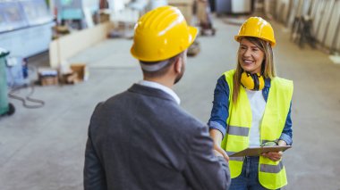 İnşaat malzemeleri ve güvenlik ekipmanı kullanan iki kişi, inşaat veya imalat alanında takım çalışması, işbirliği ve profesyonel etkileşimi simgeleyen endüstriyel bir ortamda el sıkışıyor.