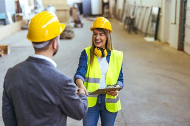 Güvenlik teçhizatı kullanan iki inşaat çalışanı, modern bir endüstriyel çalışma alanında iş birliği yaparken el sıkışırken görülüyor. Bu etkileşim, bir işyeri ortamında takım çalışması ve profesyonelliği vurgular.