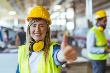 Kendine güvenen kadın inşaat işçisi güvenlik ekipmanı takıyor ve modern çalışma ortamında olumlu bir jest gösteriyor. İyimser ve profesyonel tutum takım çalışması, iş tatmini ve işyeri güvenliğine uyumu vurguluyor.