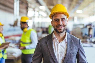 Sıkı bir şapka takan ve endüstriyel bir ortamda uygun kıyafetler giyen neşeli bir profesyonel. Meslektaşları arka planda aktif olarak takım çalışması ve güvenli, düzenli bir iş yeri ile meşgul..