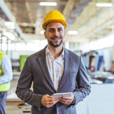 Sert şapka ve iş kıyafeti giyen kendinden emin erkek mühendis, elinde tablet tutarken fabrika ortamında duruyor, endüstri profesyonelliğini, teknik uzmanlığı ve modern işyeri teknolojisini simgeliyor..