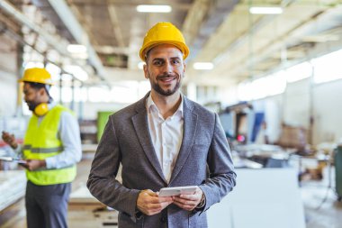 Endüstriyel bir tesiste profesyonel görünüşü olan kendine güvenen bir mühendis. Görüntü uzmanlığı, takım çalışması ve modern üretim alanındaki teknik ortamı vurguluyor.