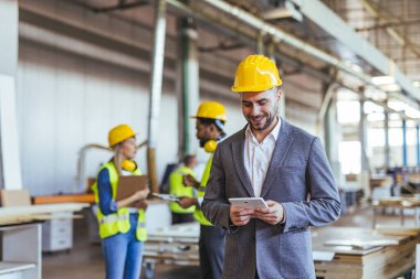 Bir fabrika ortamında, takım elbiseli ve kasklı bir adam arka planda iş arkadaşları işbirliği yaparken bir tablet kullanır, endüstriyel bir ortamda liderlik, takım çalışması, teknoloji ve üretkenlik gösterirler..