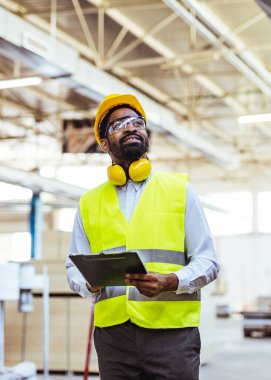 Afro-Amerikan erkek mühendis sarı kasklı, güvenlik gözlüklü ve depoda panosu olan.