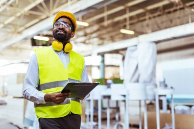 Afro-Amerikan erkek mühendis sarı kasklı, güvenlik gözlüklü ve depoda panosu olan.