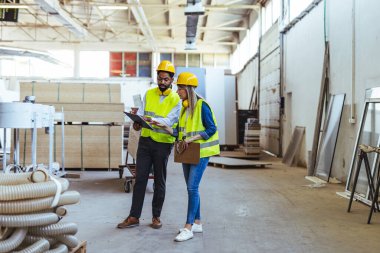 Güvenlik donanımlı iki mühendis, bir sanayi deposunu incelerken, geniş, iyi ışıklandırılmış bir çalışma ortamında üretim süreçleri ve koordinasyonuna odaklanarak bir pano üzerinde işbirliği yapıyorlar..