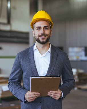 İş elbisesi ve sarı başlık giyen profesyonel bir mühendis, sanayi veya inşaat ortamında liderlik ve sorumluluk sergileyerek bir pano tutarken güven içinde duruyor. İş veya mühendislik temaları için ideal.