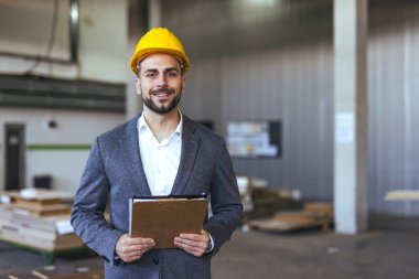 İş adamı sarı bir şapka takıyor ve elinde bir pano tutuyor. Endüstriyel bir depo ortamında geçiyor. İnşaat veya lojistik sektöründe liderlik, yönetim ve profesyonelliği temsil ediyor.