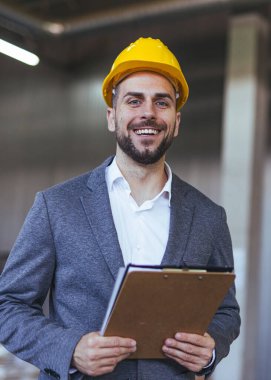 Güvenlik kaskı takmış resmi giyimli mühendis, inşaat alanında bir pano tutuyor, profesyonellik ve hazırlık yayıyor ve endüstriyel ortamdaki çalışmaları denetliyor ve denetliyor..