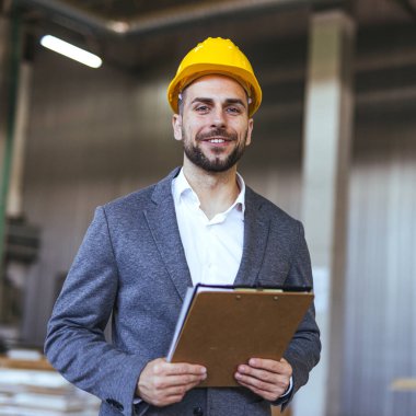 Sarı bir şapka takan gülümseyen profesyonel mühendis bir sanayi fabrikası ortamında duran bir pano tutuyor. İnşaat ve mühendislik alanlarında liderlik, uzmanlık ve profesyonellik gösteriyor.
