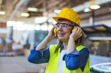 Bir fabrika ortamında, koruyucu gözlük takan ve kulaklık takan bir bayan işçi gülümsüyor. Endüstriyel bir işyerinde güvenlik, pozitiflik ve profesyonellik göstergesi