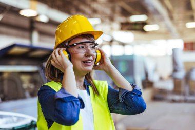 Bir fabrika ortamında, koruyucu gözlük takan ve kulaklık takan bir bayan işçi gülümsüyor. Endüstriyel bir işyerinde güvenlik, pozitiflik ve profesyonellik göstergesi