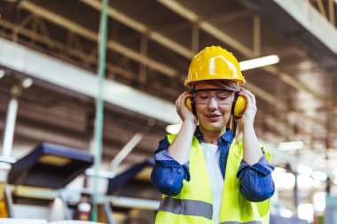 Bir fabrika ortamında, koruyucu gözlük takan ve kulaklık takan bir bayan işçi gülümsüyor. Endüstriyel bir işyerinde güvenlik, pozitiflik ve profesyonellik göstergesi