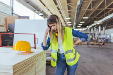 Bir inşaat işçisi bir depoda yorgun görünüyor, güvenlik giysileri giyiyor. Endüstriyel iş stresi ve depo koşullarını gösteren bir kask yakındaki bir yığının üzerine yerleştiriliyor..