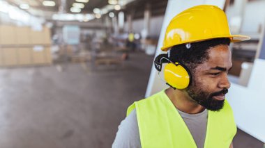 Yüksek görüşlü yelek, sarı başlık ve fabrika ortamında koruyucu kulaklık giyen odaklanmış bir işçi..