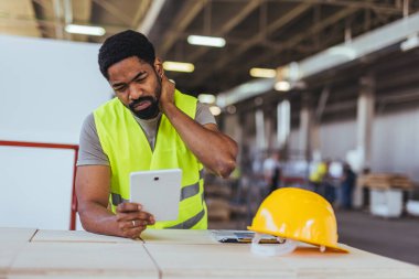 Afro-Amerikan erkek mühendis sarı kasklı ve dijital tabletli depoda boynu ağrıyan