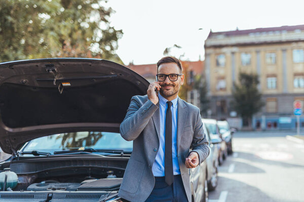 Бизнесмен в костюме, используя свой мобильный телефон рядом с машиной с открытым капюшоном, указывает на поломку, будучи спокойным и находчивым, призывая к придорожной помощи в городской среде.
