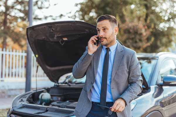 A businessman in formal attire is stranded beside his car with the hood open, making a phone call for assistance, conveying urgency and a professional scenario in an outdoor setting.