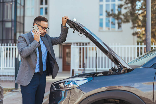 A professional man in formal attire examines a car with the hood open while using his phone, illustrating mechanical issues and troubleshooting in a modern urban context.