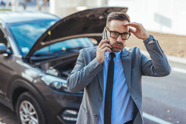 Профессионально одетый человек звонит за помощью возле своего автомобиля-инвалида с капюшоном, отражая момент беспокойства и практического решения проблем в городской обстановке..