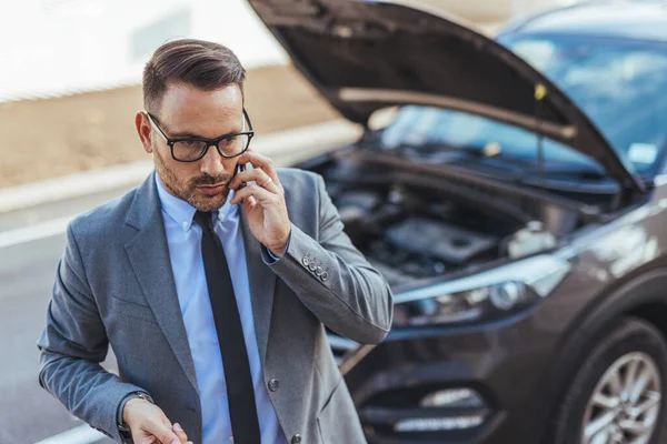 Profesyonel giyimli bir adam yol kenarında duran arabasının yanında bir telefon görüşmesi yapar. İş ortamında acil durum ve sorun çözme işlemleri yapar..