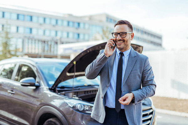 A smiling businessman in formal attire using a cellphone while standing beside a broken-down vehicle. The car's hood is open, indicating a mechanical problem. Urban building background adds to context.