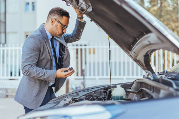 A businessman in a suit inspects a car's engine outdoors, focused on checking the vehicle's condition. The image captures attention to detail, responsibility, and the examination of technical issues.