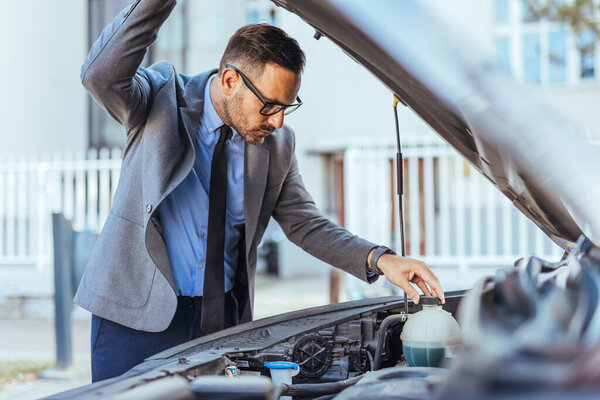 A professionally dressed businessman inspects a car engine outdoors, expressing concern and curiosity. The urban setting and formal attire suggest an unexpected issue. Ideal for concepts of troubleshooting, vehicle issues, and problem-solving.