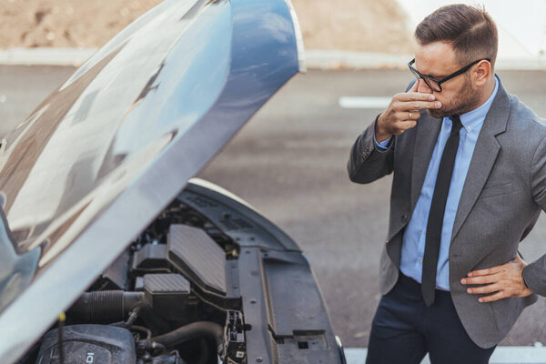 A formally dressed man inspecting a car's engine while displaying concern, possibly addressing a mechanical problem. The casual environment contrasts with the formal attire, emphasizing adaptability and problem-solving skills.