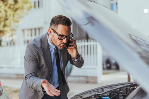 A professional man wearing a suit is on a phone call examining a car with an open hood, appearing concerned.