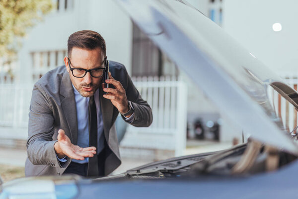 Профессиональный бизнесмен в костюме осматривает свой двигатель автомобиля, обсуждая проблему на телефоне. Изображение отражает озабоченность и прерывание повседневной жизни из-за проблем с транспортным средством.