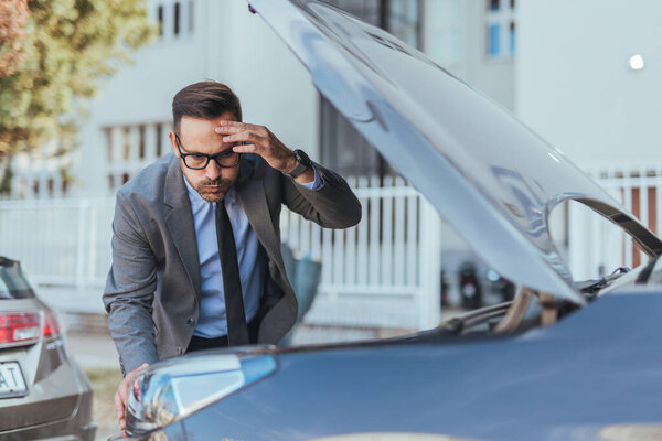A serious businessman wearing a suit inspects a car's damaged engine in an urban setting, seemingly encountering mechanical trouble. His concerned expression highlights technical issues and roadside emergencies.
